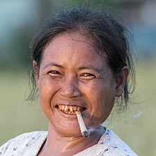 Portrait d'une femme serrant une cigarette roulée à la main entre ses dents, au coucher du soleil dans la campagne de Don Det, Laos. La couleur rouge de ses dents est due à la mastication régulière de paan. Septembre 2017.