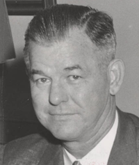 headshot of a middle aged white man with a tight haircut. He is wearing a suit jacket and a tie