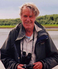 Colour photograph of Stan Paterson standing, with a camera in his hands and binoculars around his neck