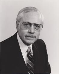 Photo of Hal Trumble wearing a dark jacket, white shirt, striped neck tie, and tinted glasses.