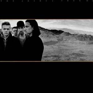 A landscape monochrome photograph of U2 in the desert sits in the center of a black background. U2 are standing on the left half of the photograph, with a mountain range on the right half. Tiny gold text reading "THE JOSHUA TREE U2" is stretched across the top of the black background.