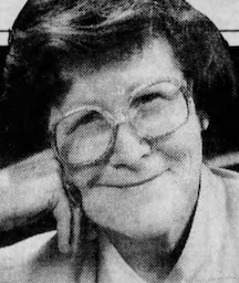 An older white woman, smiling, wearing glasses; her head is resting on her folded right hand.