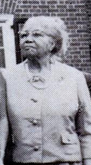 An elderly black woman, wearing a light-colored suit and glasses.
