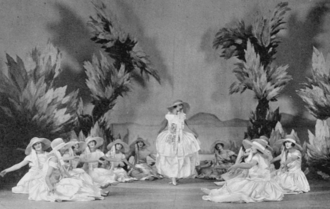 A young white woman standing, with female chorus seated in a circle on the ground around her