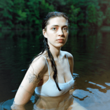 A wet woman wearing a white undershirt stands half submerged in a natural body of water in the woods. Her nonexpressive face looks at the camera.