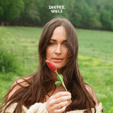 A woman with very long brunet hair holds up a flower against a bucolic green backdrop.