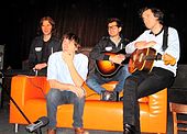 Четверо мужчин сидят на оранжевом диване, двое держат гитары, а один за клавишными, установленными на левом подлокотнике дивана.