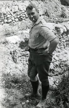 A middle-aged man in shirt sleeves and trousers, his hands on his hips, smoking a cigarette.