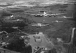 Head-on view of seven four-engined military aircraft flying over an airfield