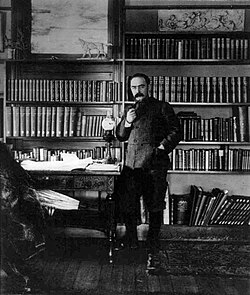 Full length photo of a man dressed in a khaki jacket standing in a book lined study. He has short hair, a moustache and is smoking a pipe.