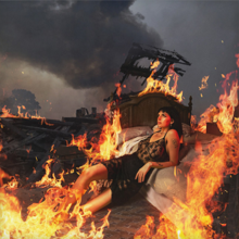 Black reclining against a burning bed in a fiery outdoors scene.