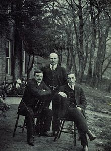 Three men in dark suits, two sitting on chairs and the third stood behind