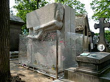 A large, rectangular, granite tomb. A large, stylised angel, leaning forward is carved into the top half of the front. There are a few flowers beside a small plaque at the base. The tomb is surrounded by a protective glass barrier that is covered with graffiti.