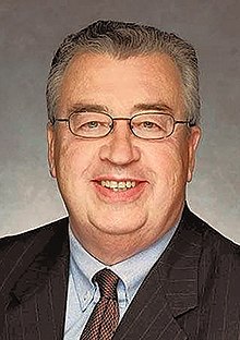 An older man with thin gray hair and glasses smiling, wearing a jacket and tie, against a gray background