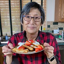 A woman wearing glasses and a checked red and black shirt holds a plate of waffles with strawberries, whipped cream, syrup, and powdered sugar