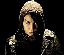 A young woman in a hooded black leather jacket and spiked collar necklace, with heavy eye-makeup and black lipstick on, and piercings in her eyebrow and nose, looks coldly into the camera.