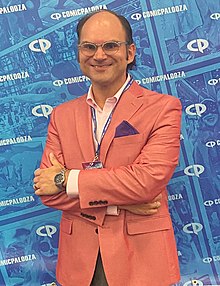 An olive-skinned Jewish man with long black hair that is bald on top. He is wearing translucent browline eyeglasses. He is wearing a salmon-colored blazer and open collar pink shirt with a purple pocket square. His arms are crossed and a silver chronograph watch is visible on his left wrist.