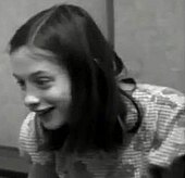 Черно-белый кадр из видео, на котором Джини, восторженно улыбается, снято с расстояния в несколько футов. Она показывает ее от груди вверх, а Джин смотрит чуть правее камеры.