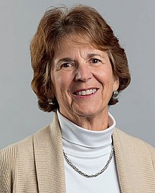 Portrait of a white woman, around sixty years of age, with brown hair, facing the camera. She wears a beige jacket and an off-white turtleneck.
