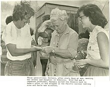 An older white woman with white hair, center, greets a younger white woman, left, while a teenaged girl with dark hair and glasses looks on, right.