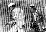Black-and-white image of two men facing the left of frame, walking in front of a brick wall. A bold series of vertically striped shadows covers the entire image. The middle-aged man to the right wears a white fedora, a medium-dark suit, and an open-collared white shirt. In front of him, to the left of the image, a younger, taller man wears a cream-toned suit, a white beret and shirt, and a light striped tie. Each man holds a pistol in his right hand.