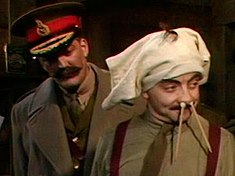 A man with a large moustache wearing a general's cap stands behind a man wearing underpants on his head and with pencils in his nose.