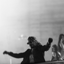 Future with bleached blonde dreadlocks, wearing sunglasses and a black jacket, throws his hands in the air, letting his jacket flow in the wind, while standing in front of a black-and-white foggy background. Three women can be seen standing in the background.