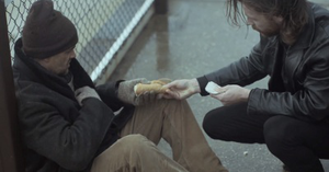 A screenshot from the music video; Browne's character is giving a homeless man (portrayed by Carter) food and taking back his money he offered him first. The video has low colouring and is set on a dreary day, puddles are still present from the rain.