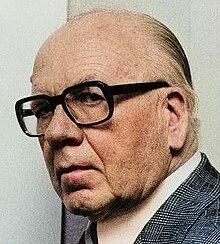 Three-quarter view of elderly man's face: clean-shaven, with dark-rimmed spectacles and receding hair.