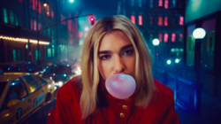 Dua Lipa blowing a bubble while sitting on a car in a traffic jammed city block.