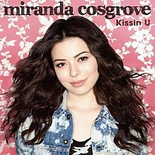 A brunette woman standing in front of a pink and white background. She wears a bleached-out demin jacket and a black and white shirt. The words "Miranda Cosgrove" (stylized in lowercase letters) and "Kissin U" are written above her.