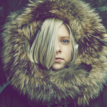 A blonde woman with a brown parka is staring forward against a forest background. The subject's hair covers the right side of her face.