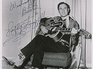 A dark-haired man wearing a neckerchief, a shirt with fringes dangling from the sleeves, and pants with a guitar pictured on them, smiling broadly while leaning on a guitar