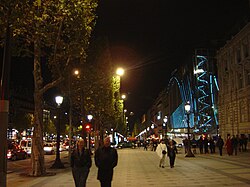 The Champs-Élysées (seen from the Place de l'Étoile) are busy even in the late evening; cinemas, night clubs and restaurants attract a clientele. On the right, the Drugstore Publicis, open late, sells many wares, including upscale take-away food.