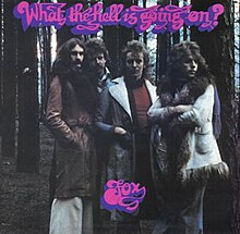 A washed out colour photograph with purple scrawled lettering What The Hell Is Going On? along the top and Fox the band name on the bottom center. The image is the four members of the band to the knee facing the viewer against the backdrop of straight dark forest of conifer tree trunks.