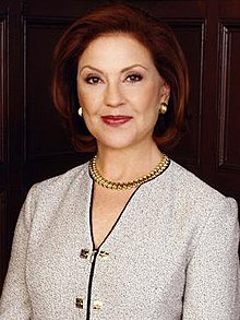 Elderly woman with short auburn hair smiling and dressed in upper-class attire, including a pair of pearl earrings, a gold pearl necklace and light purple blazer.