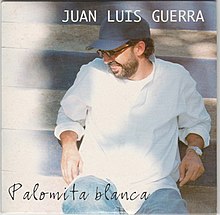 Image of Juan Luis Guerra wearing a white shirt, dark blue cap, sitting down and facing right.