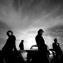 A black-and-white photo of four men leaning against different sides of a convertible car with a cloudy sky behind them.