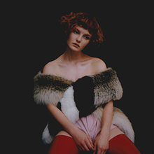 A young woman with red hair wearing fur sitting and staring into the distance in front of a black background.