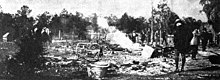 =A photograph of ashes from a burned building with several people standing nearby and trees in the distance