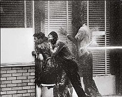 Three black high school students, two boys and a girl, facing into a storefront window to avoid being hurt by a water cannon striking one boy at his back; all three are dripping with water
