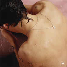 Harry Styles is half submerged in a pastel pink bath, both his hands cover his face while his back faces the viewer. Around his neck are two silver necklaces, with one of them flipped to his back to show a tiny lotus flower pendant.