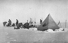 Six men are working with sleds and camping equipment, close to a pointed tent pitched on a snowy surface. Nearby, upright skis have been parked in the snow