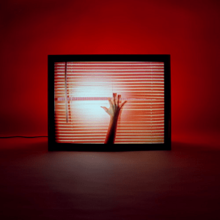 A dark room with red light glowing, a TV is plugged in with a hand touching over window blinds.