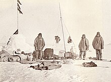 Слева - снежная пирамида с флагами. Рядом трое мужчин, а на снегу разбросано разное снаряжение.