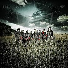 A group of nine men wearing masks stand in a field of grass with a gloomy clouded sky above them.