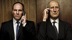 Head and shoulders of two serious-looking men in dark suits standing side-by-side, facing the camera, in front of dark wooden panels, each holding a phone to their ear.