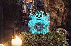A blue furry toy with a flower crown sitting on a shrine.