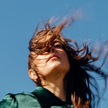 A photograph of Madi Diaz, a light-skinned woman, wearing a turquoise jacket, with her hair being swept by the wind across her face, in front of a blue background.