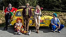 Four clowns and one woman lean against a yellow clown car